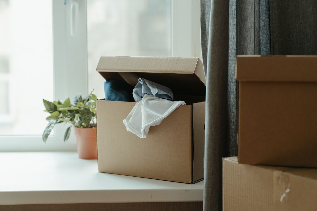 our-story Open cardboard box with clothes by window, symbolizing moving or relocation.